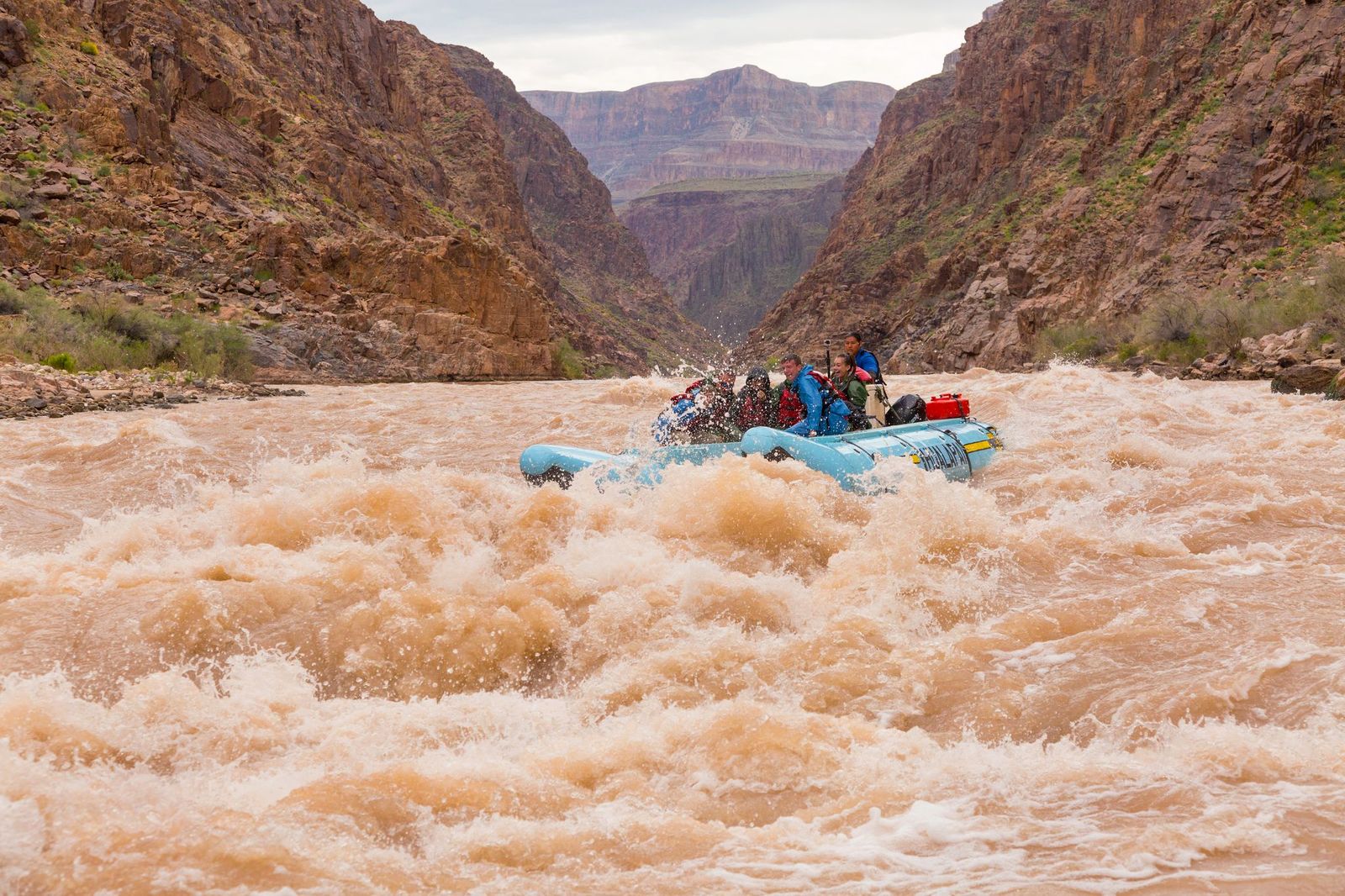 A raft carrying several people navigates turbulent, muddy brown rapids on a river in a deep canyon surrounded by towering, reddish-brown rock walls.