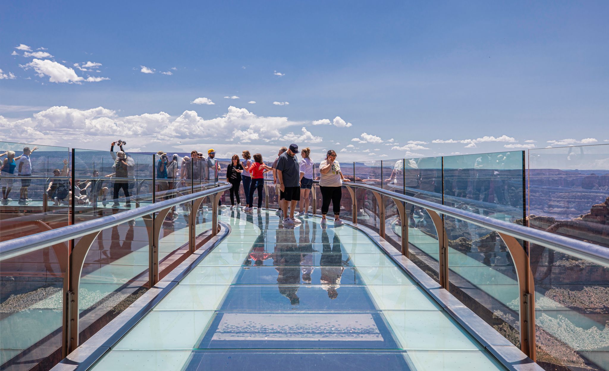 
A group of tourists is walking on the U-shaped, glass-floored Skywalk over the edge of the Grand Canyon, which offers panoramic views of the distant canyon landscape under a bright blue, cloudy sky.