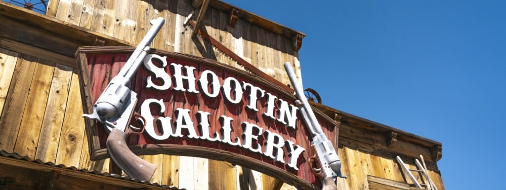 A "Shootin' Gallery" sign with two cowboy revolvers hangs on a wooden building.