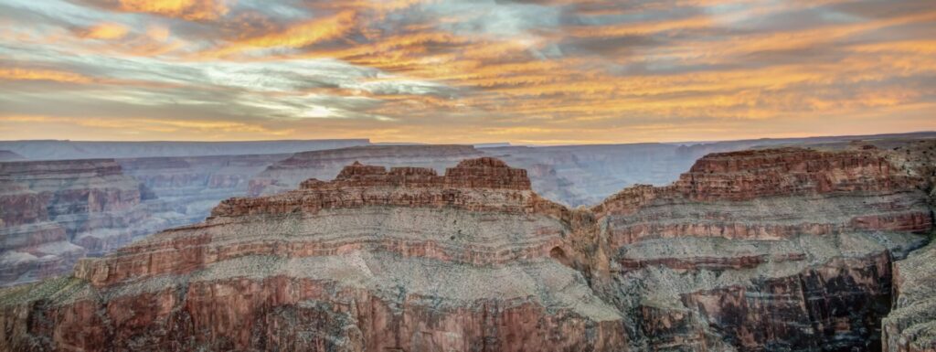 A dramatic sunset or sunrise sky with orange and yellow clouds hangs over the vast, layered Grand Canyon.