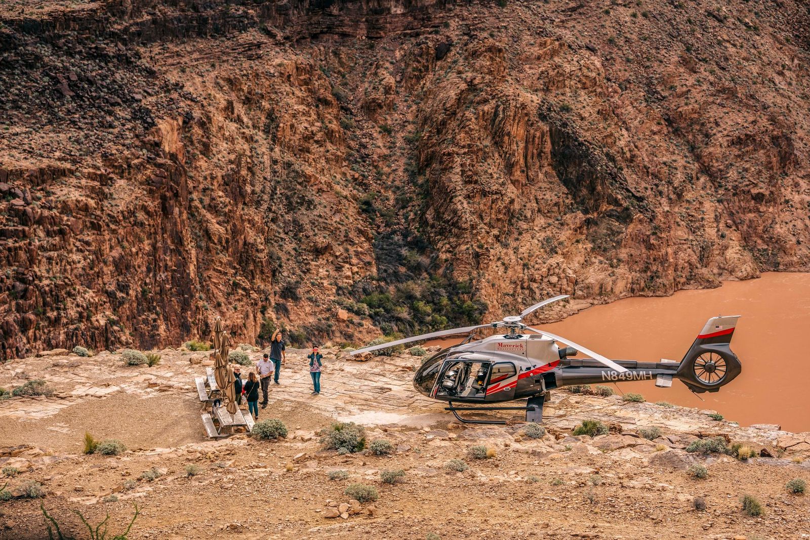 A gray and red Maverick helicopter is landed on a dusty, rocky ledge next to a muddy, orange-brown river, while a small group of tourists walks nearby against a backdrop of steep, reddish-brown canyon walls.