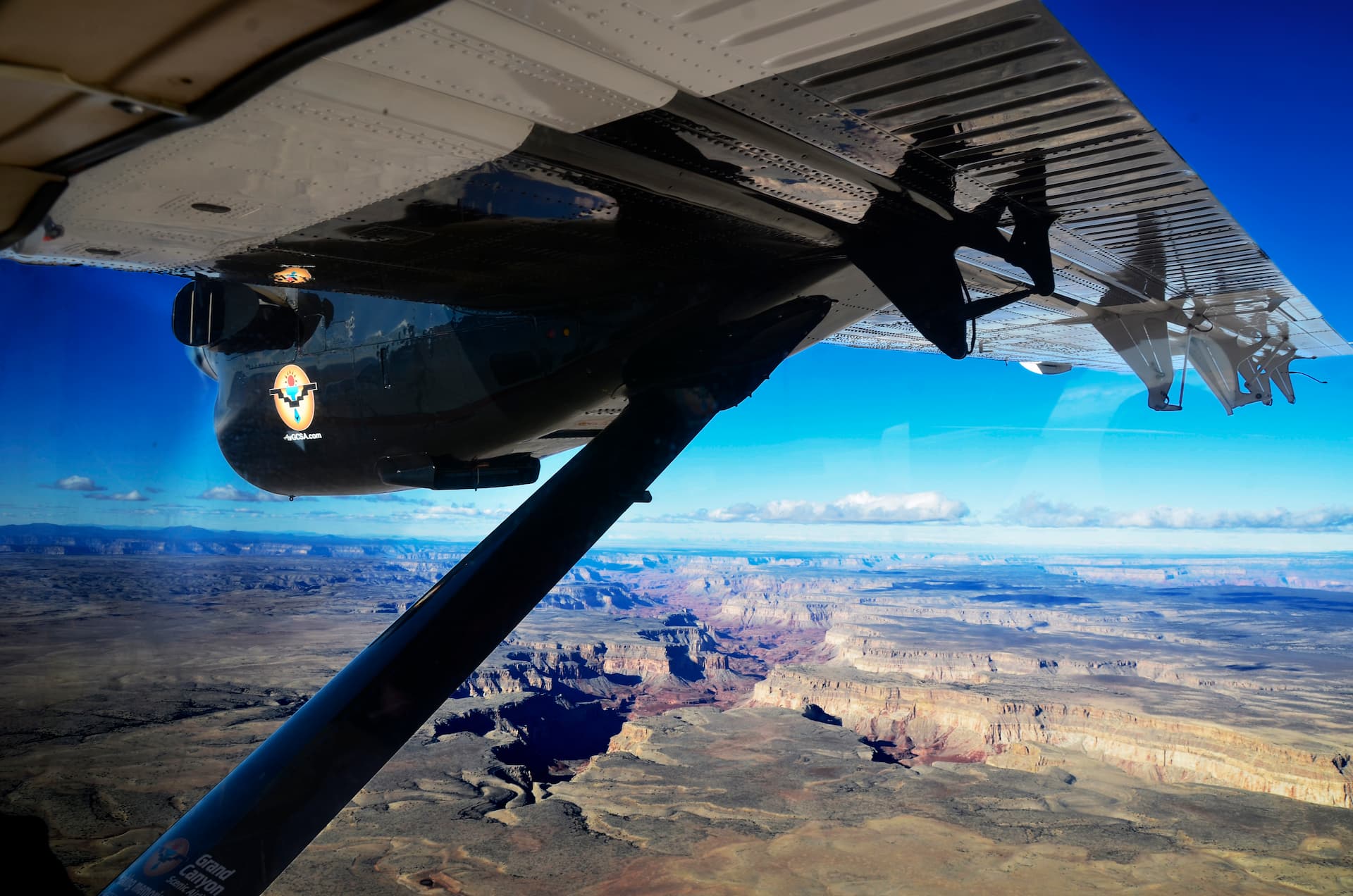 An aerial view of the Grand Canyon taken from the window of a sightseeing airplane. The plane's wing is visible in the foreground, with the vast, layered canyon landscape unfolding below under a deep blue sky.