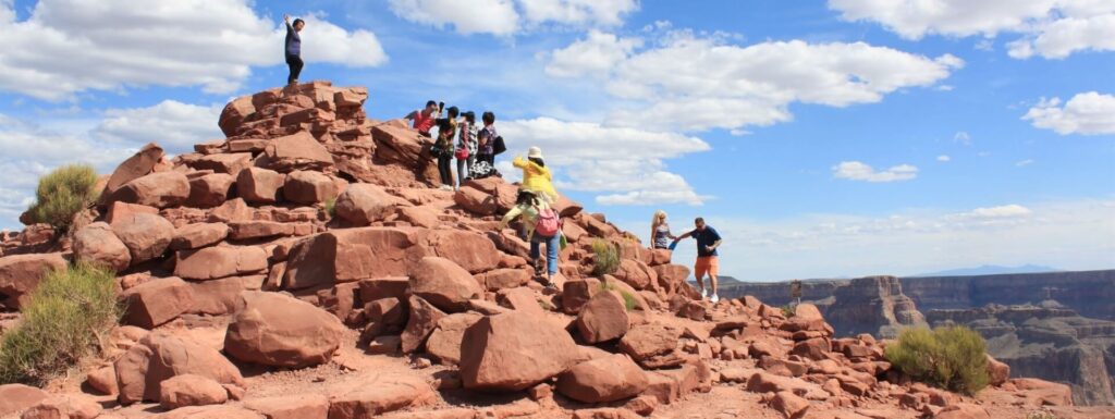 People are climbing and standing on a rocky hill overlooking the Grand Canyon.