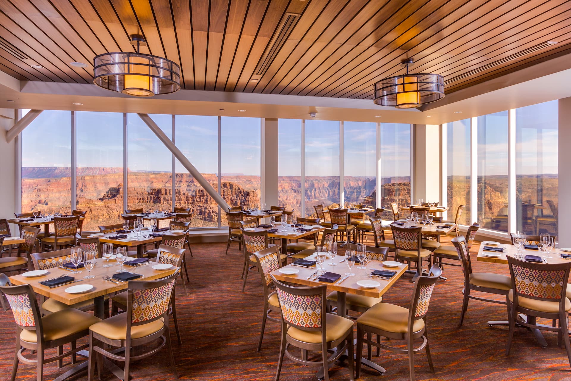 The interior of a restaurant with floor-to-ceiling windows that offer a stunning panoramic view of the Grand Canyon. The dining room is elegantly set with tables and chairs, awaiting guests.