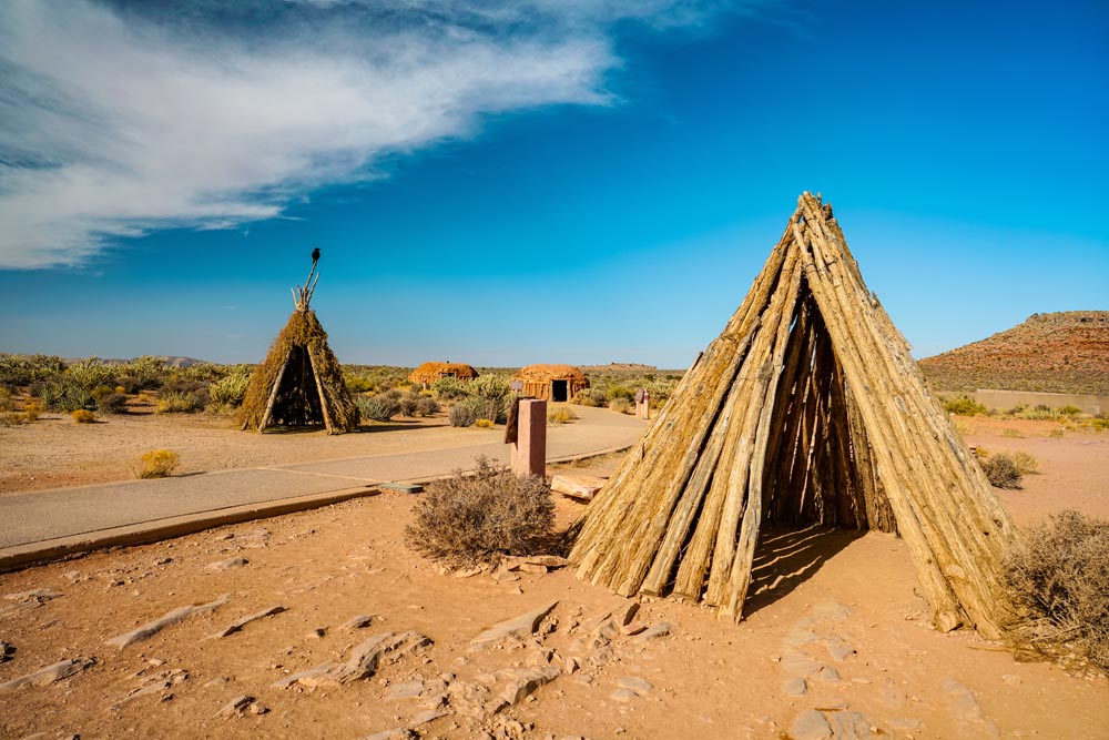 Two teepee-like structures made of logs stand in a desert landscape under a blue sky.