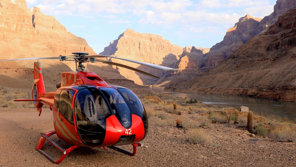 A red helicopter is parked on a dirt path next to a river in the Grand Canyon.