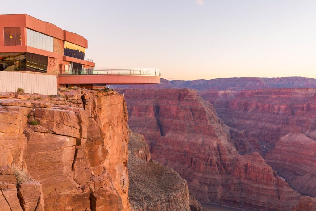 A glass skywalk extends from a modern building over the Grand Canyon at sunset or sunrise.