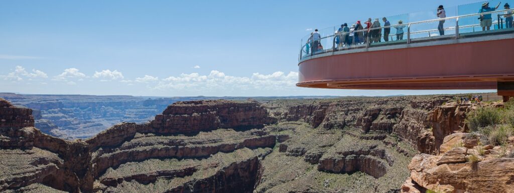 A panoramic view of the Grand Canyon shows the Skywalk on the right, with people looking out over the vast landscape. 