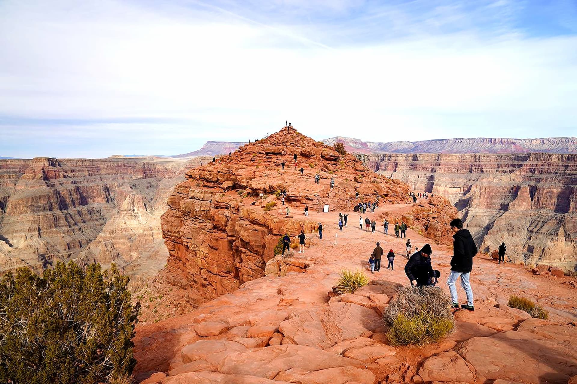 Tourists explore Guano Point, a large red rock viewpoint that extends into the Grand Canyon. The vast, layered canyon walls stretch into the distance under a bright sky.