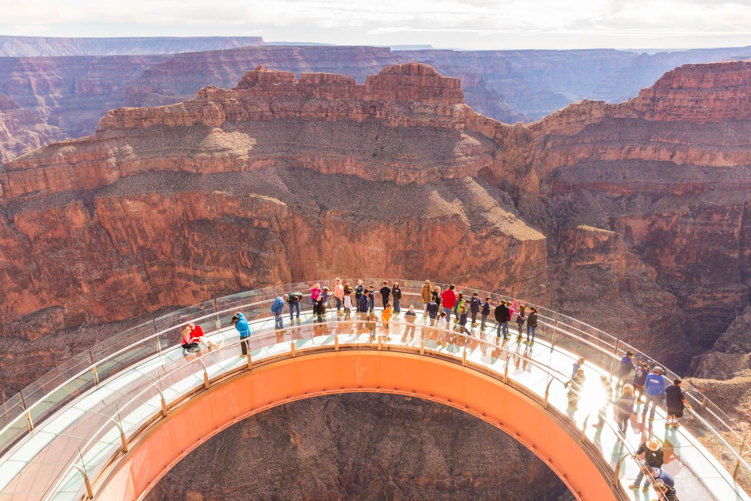 A high-angle view of tourists standing on the Grand Canyon Skywalk, a large, horseshoe-shaped glass bridge extending over the vast, layered red rocks of the canyon.
