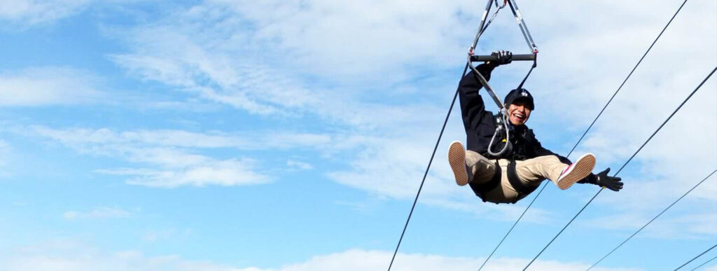 A person smiles while zip-lining against a blue, cloudy sky.